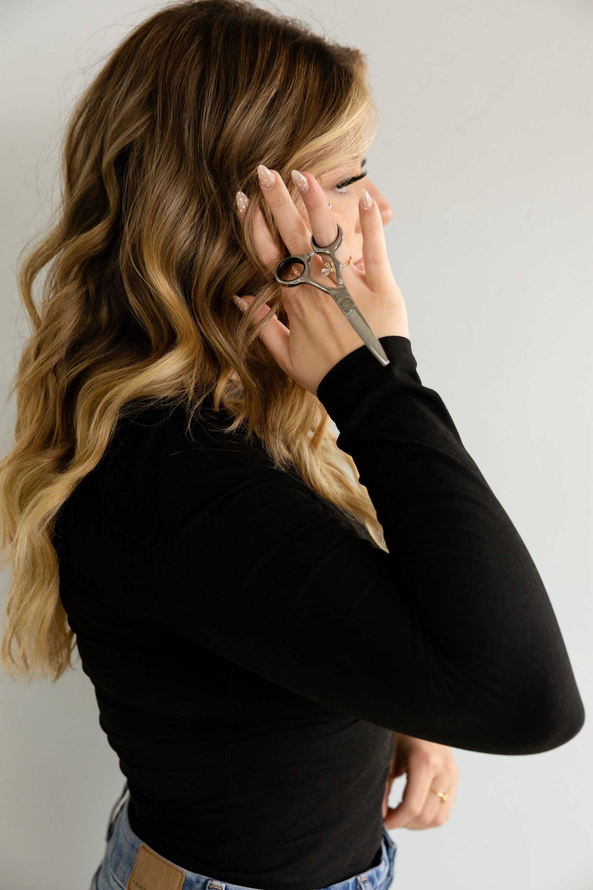Woman with long wavy hair holding hair scissors near her head, wearing a black top.