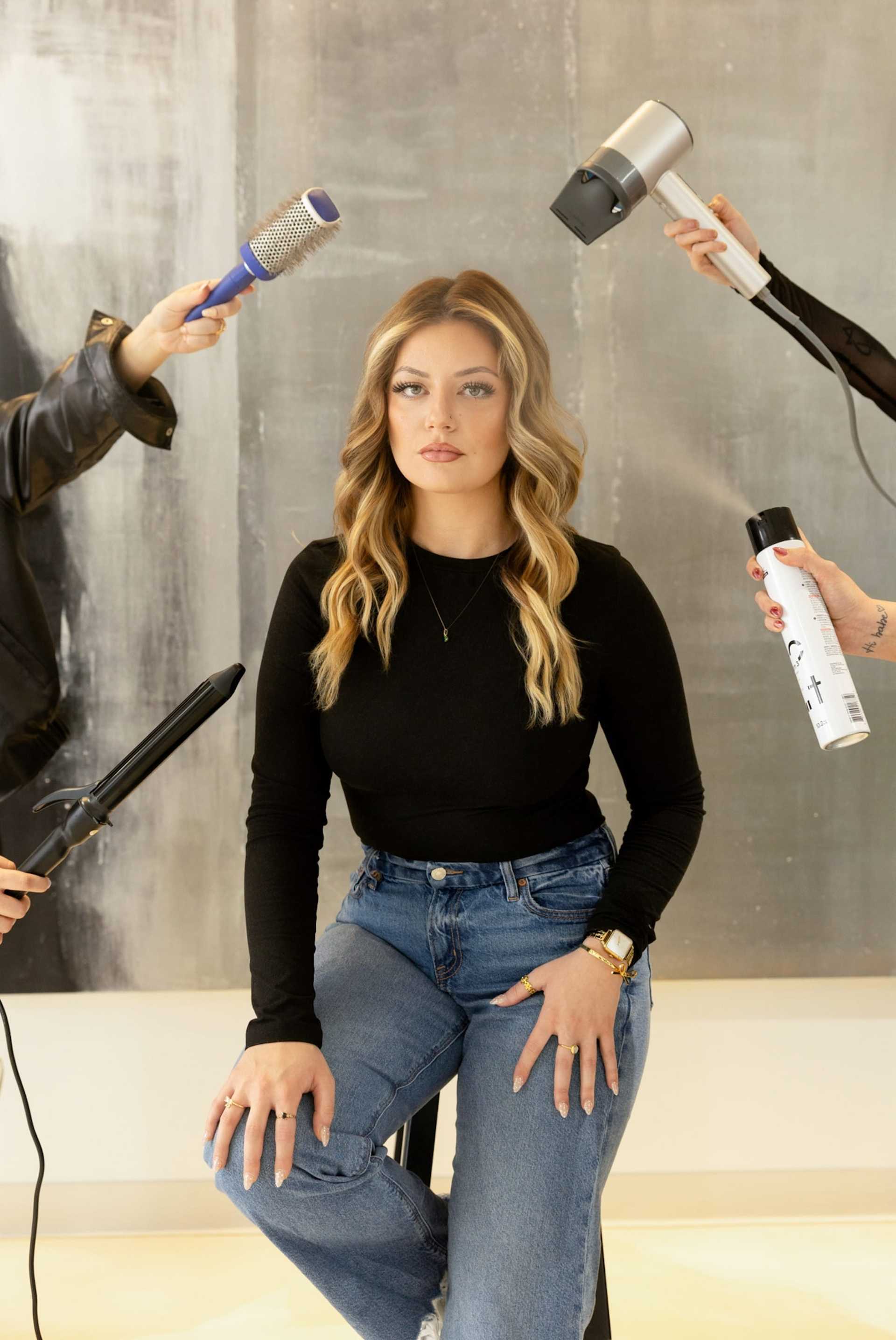 Young woman sitting while hairstylist holds tools around her for styling.
