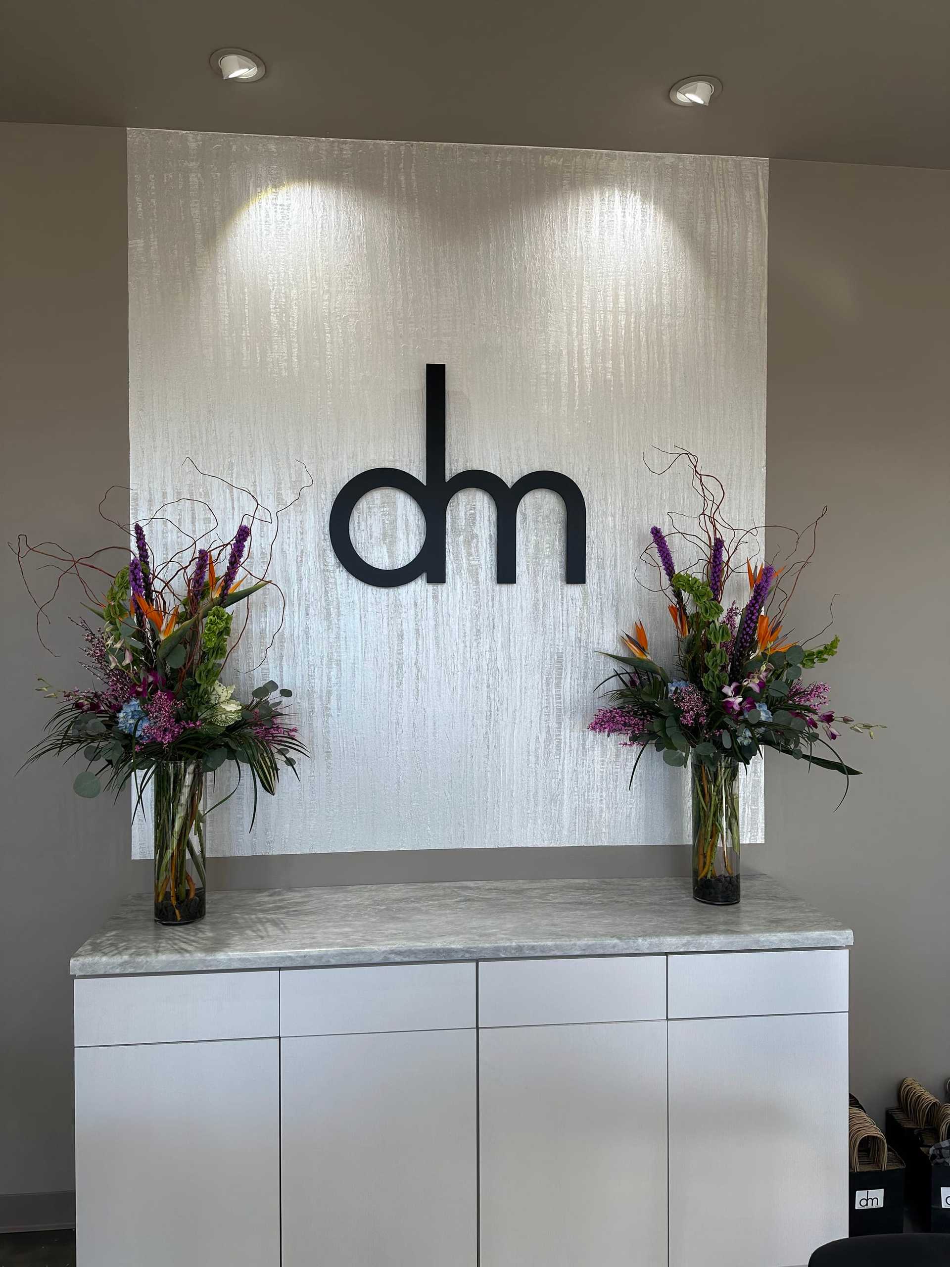 ```plaintext
Modern lobby with 'dm' logo and floral arrangements on a sleek white cabinet.
```