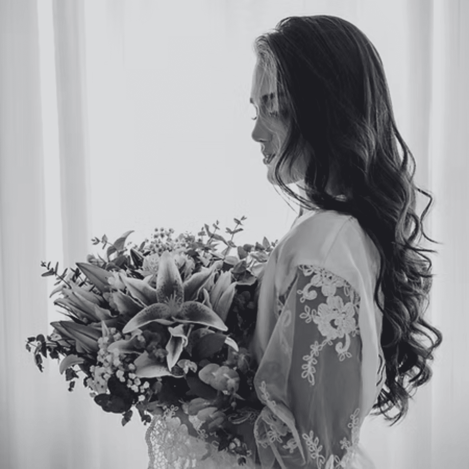 Bride holding bouquet, captured in profile, wearing lace robe against soft light background.